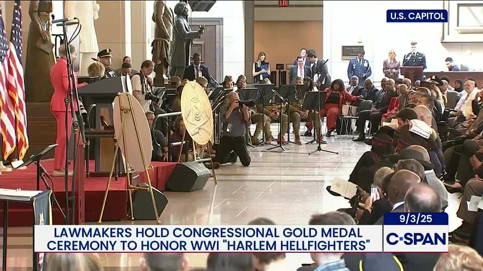 A woman in a red suit speaks at a podium in front of a stage, with the American flag to her left. Photographers crouch and stand, capturing the event for C-SPAN.
