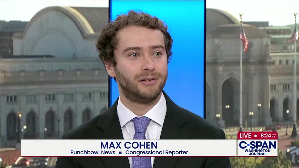 A reporter, Max Cohen, is speaking on C-SPAN's "Washington Journal" broadcast. Behind him, the backdrop shows a stately building, and American flags are waving in the breeze.
