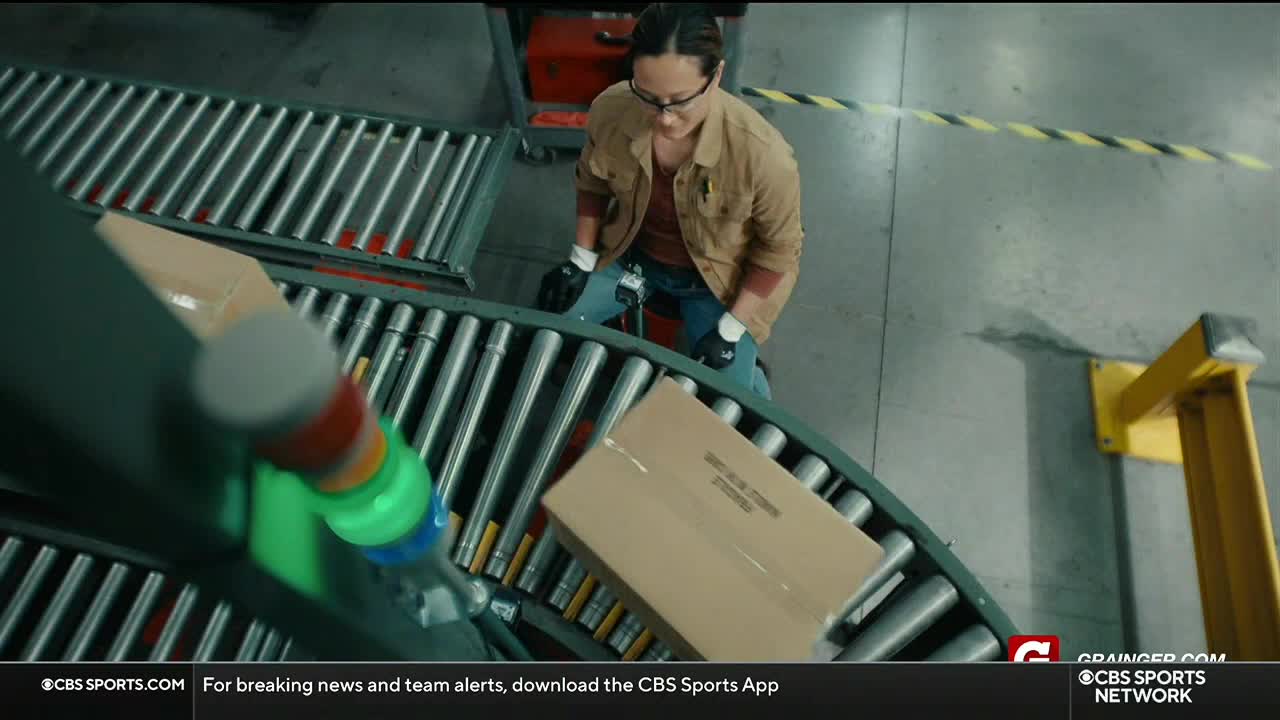 A woman in a brown jacket and glasses is carefully guiding a cardboard box along a roller conveyor. The CBS Sports Network USA logo is visible in the corner.
