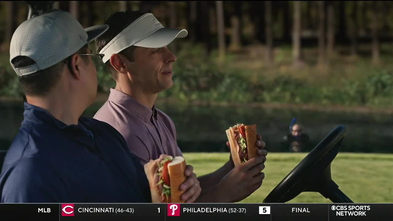 Two men in golf attire sit in a cart, each holding a sandwich with visible lettuce and tomatoes. The CBS Sports Network USA logo is in the corner, and the score of a baseball game is displayed at the bottom.
