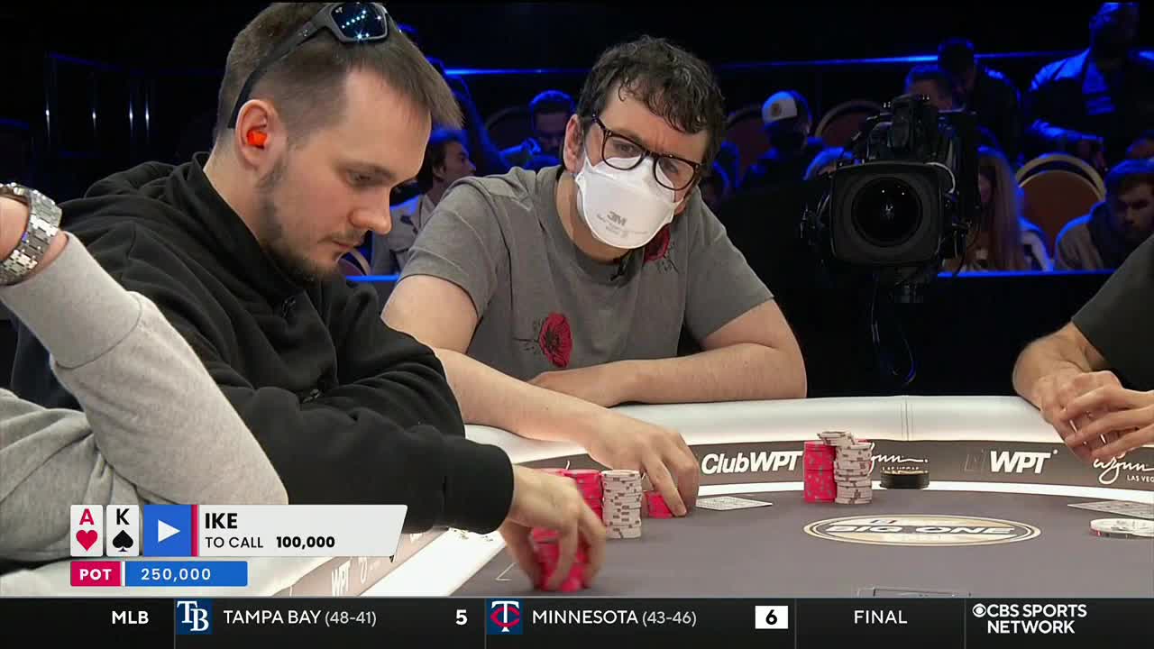At a poker table, a player in a gray t-shirt and a mask is watching intently, while another, wearing sunglasses atop his head, is focused on the cards. The CBS Sports Network USA graphic at the bottom shows the score of a baseball game.
