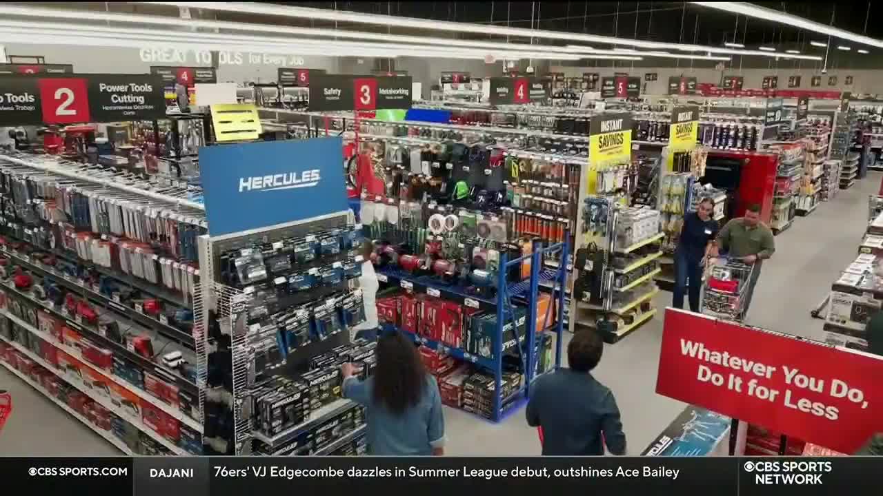 Shoppers browse the aisles of a hardware store, with a couple pushing a cart down the center. The CBS Sports Network logo is visible in the bottom corner.
