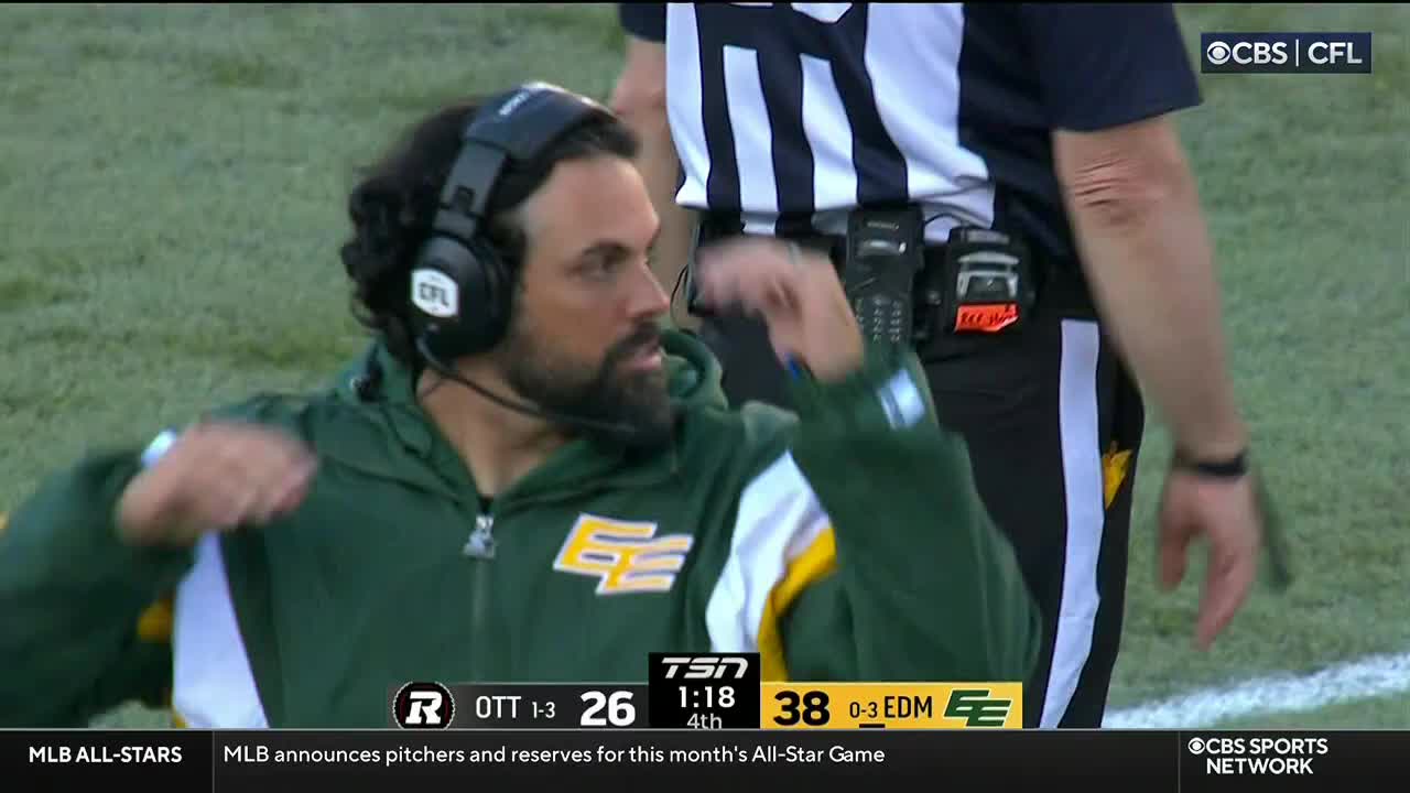 On the sidelines during a CBS Sports Network USA broadcast, a coach in a green jacket gestures emphatically, wearing a headset. Beside him, a referee in black and white stripes stands still, watching the game.
