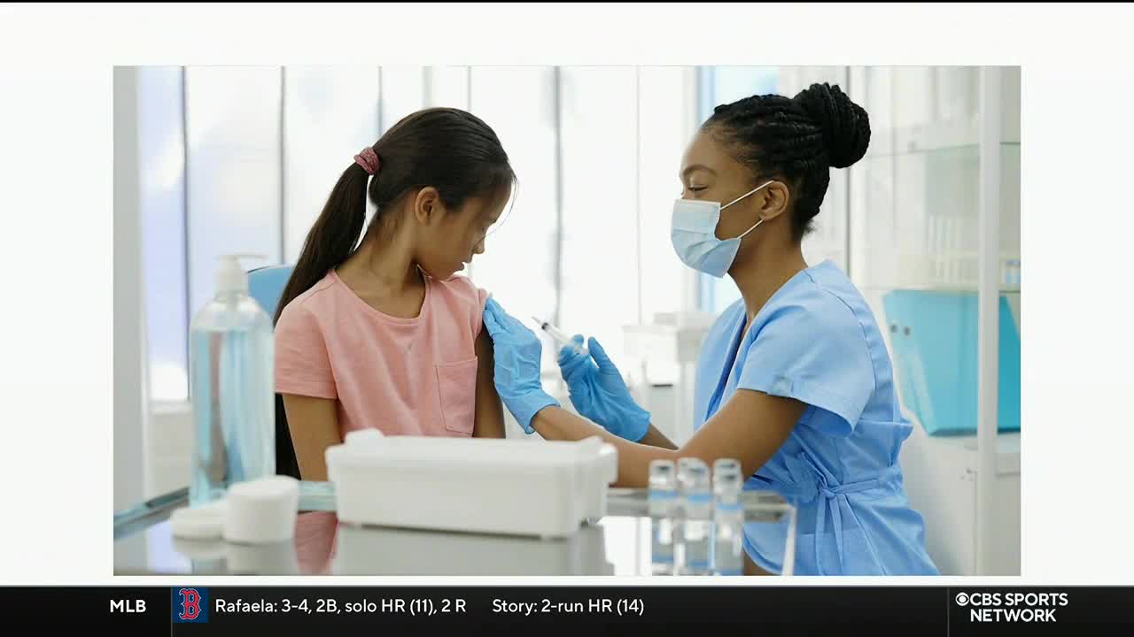 A nurse in blue scrubs is injecting a vaccine into a young girl's arm. The child is looking down, and the needle is just entering her skin.
