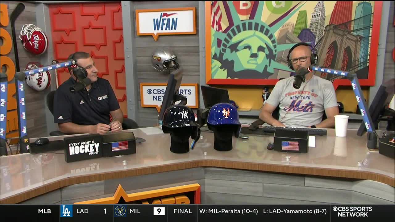 Two men sit at a desk, each with a microphone in front of them. In the foreground, baseball helmets sit between them, with a "CBS Sports Network" logo visible in the background.
