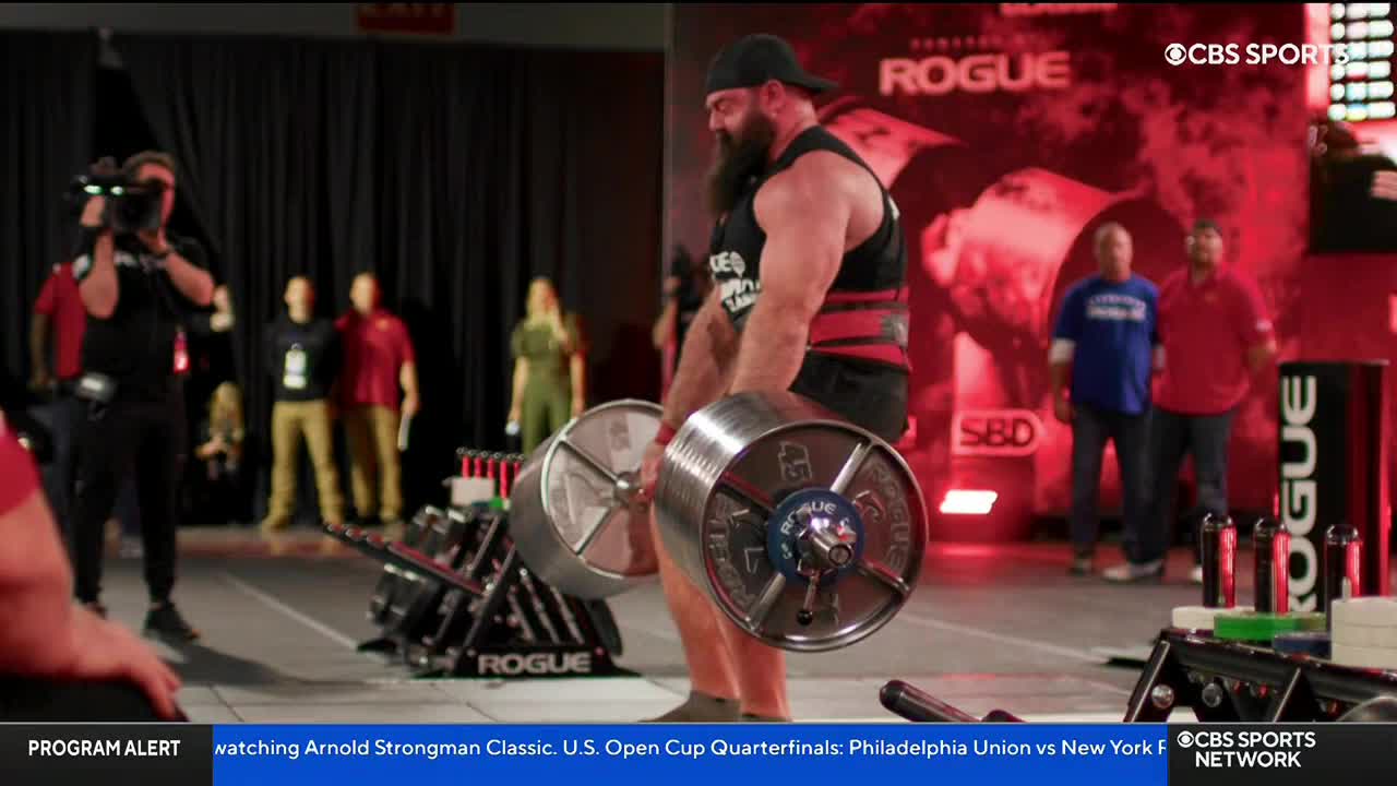 A competitor, wearing a black tank and a red weightlifting belt, strains to lift a heavy barbell. The event is being broadcast on CBS Sports Network USA, and the crowd watches intently.
