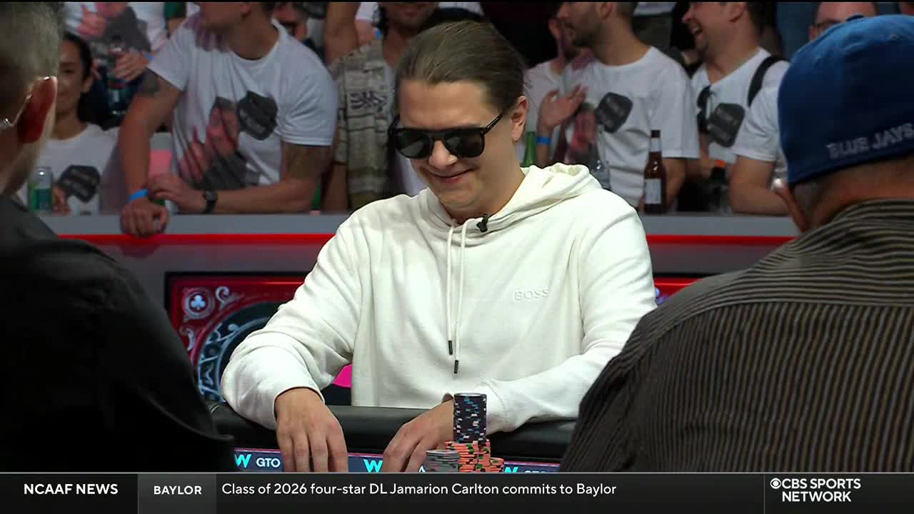 A man in a white hoodie and sunglasses smiles slightly, looking down at the poker table. In the background, a crowd watches the game, visible on CBS Sports Network USA.
