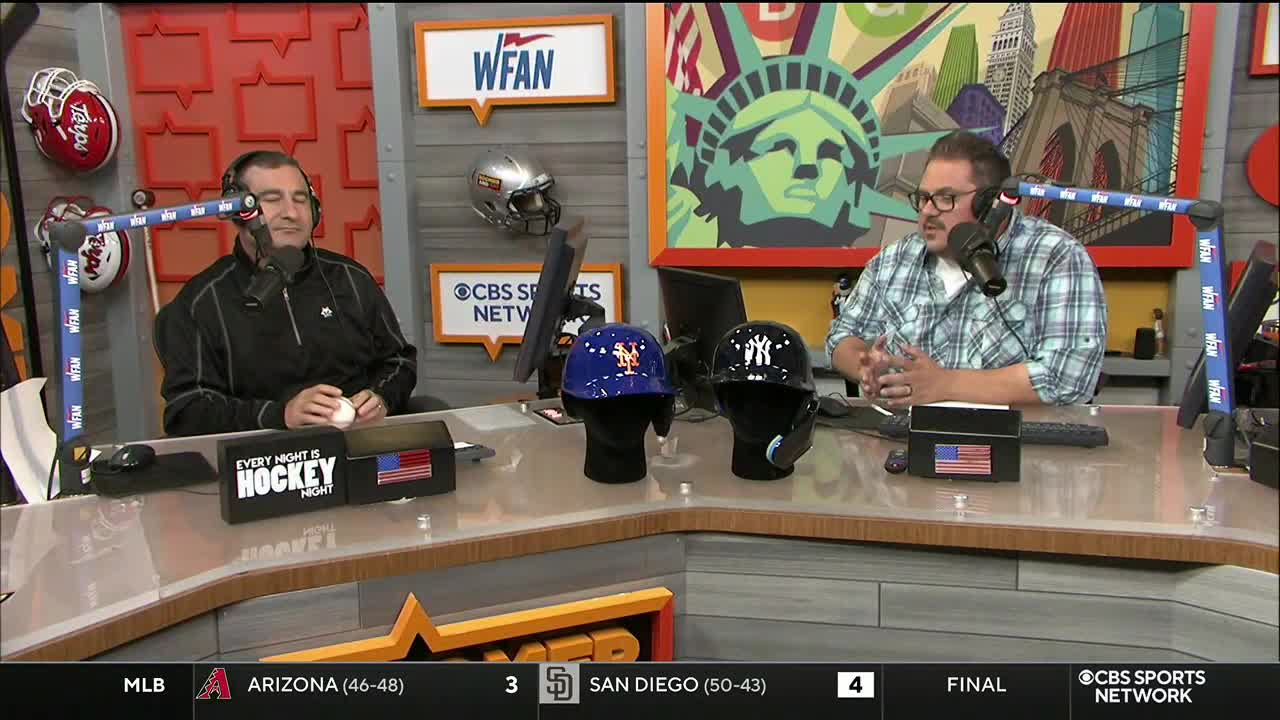 Two men are seated at a desk, each wearing a headset and speaking into a microphone. Between them are two baseball helmets, one blue and one black, with the logos of the New York Mets and Yankees clearly visible.
