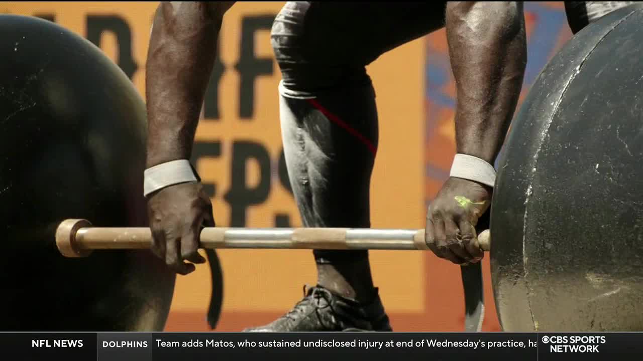 A strongman, competing on CBS Sports Network USA, grips a barbell, his knuckles white and veins bulging. He's about to lift, the massive black spheres flanking him.
