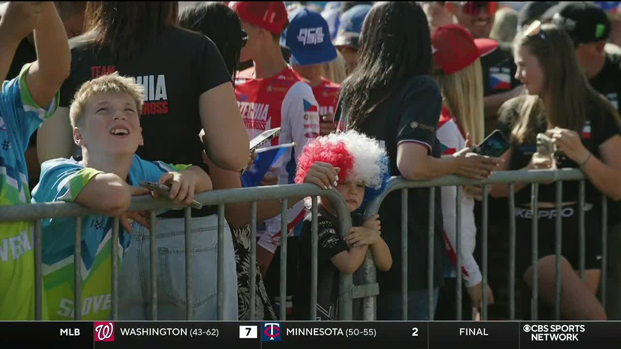 A young boy in a blue and green shirt leans on the barrier, gazing upwards with a look of excitement. Beside him, a child with a red, white, and blue wig clings to the fence, watching the action.
