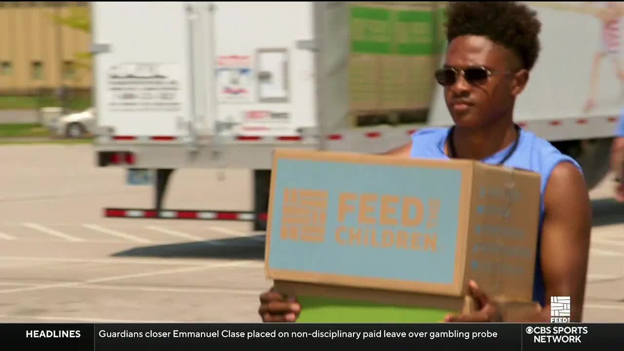 A young man, wearing sunglasses and a blue shirt, carries a cardboard box labeled "FEED! CHILDREN." Behind him, a semi-truck trailer is parked, and the scene is broadcast on CBS Sports Network USA.
