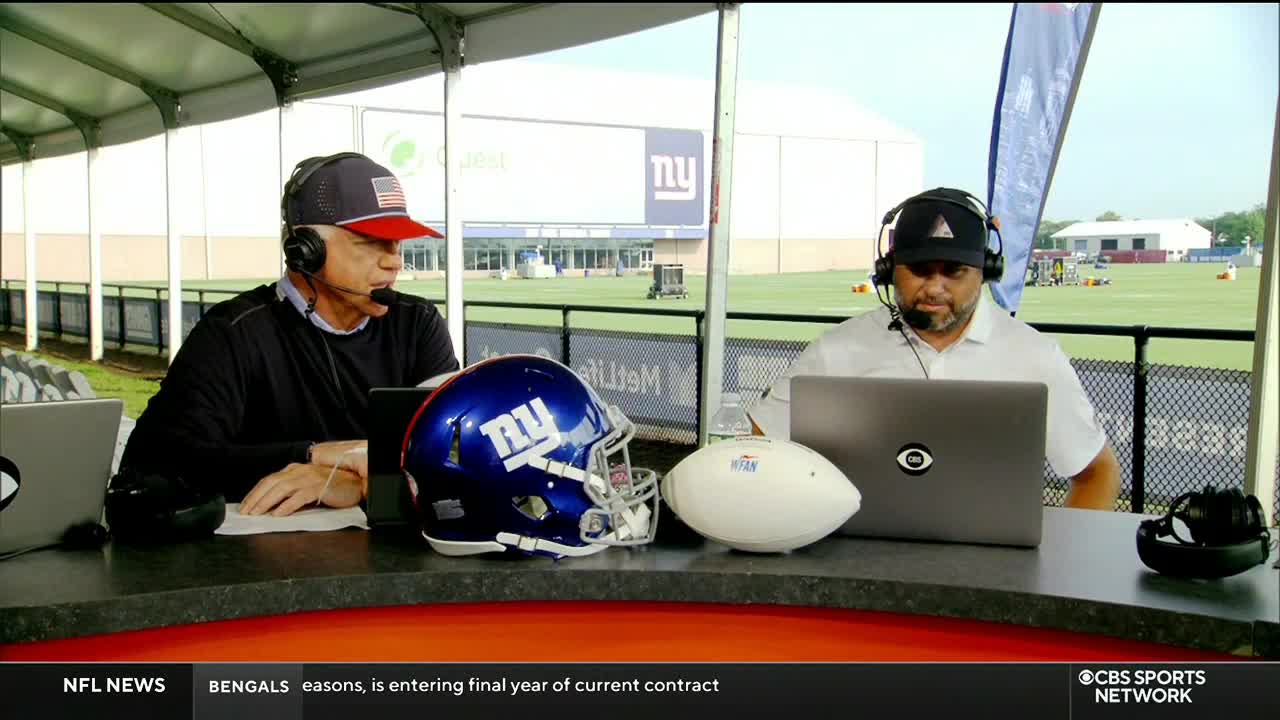 Two men in headsets sit at a table, a New York Giants helmet and a football between them. They're broadcasting from a tent, with the practice field visible behind them on CBS Sports Network USA.
