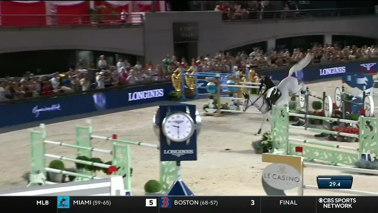 A white horse and rider are soaring over a green and white fence, their bodies angled in mid-air. The crowd in the stands watches as the horse clears the jump, the CBS Sports Network USA logo visible in the corner.
