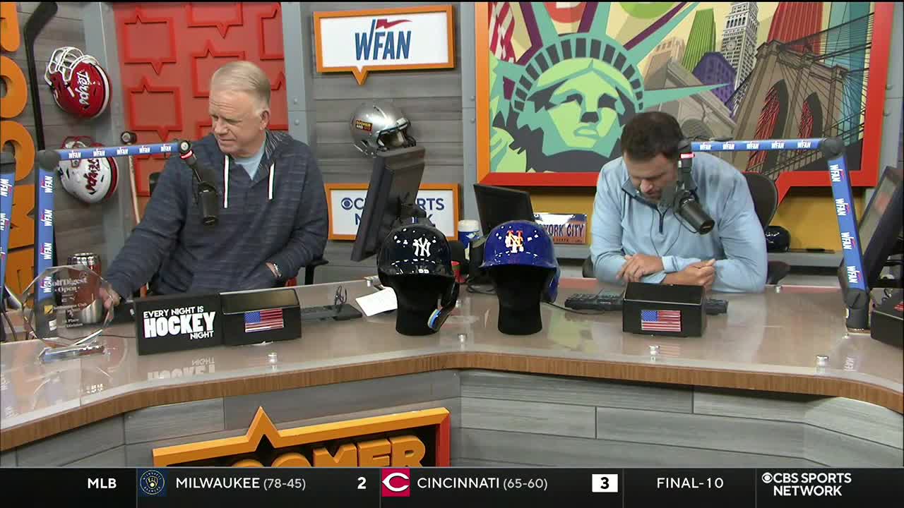 Two men sit at a desk on a CBS Sports Network USA set, each with a microphone in front of them. Between them, two baseball helmets sit on display.
