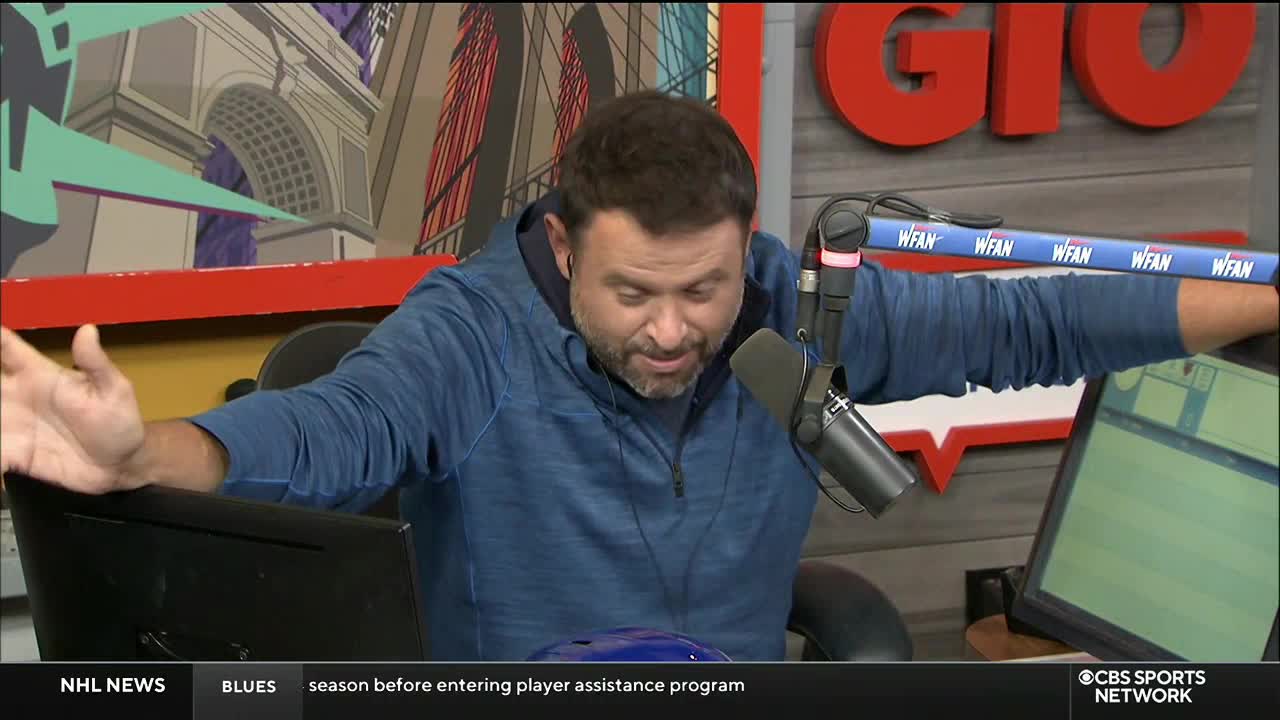 The man in the blue hoodie has his arms outstretched, seemingly gesturing to something just out of view. His face is turned down, and he appears to be speaking into a microphone from CBS Sports Network USA.

