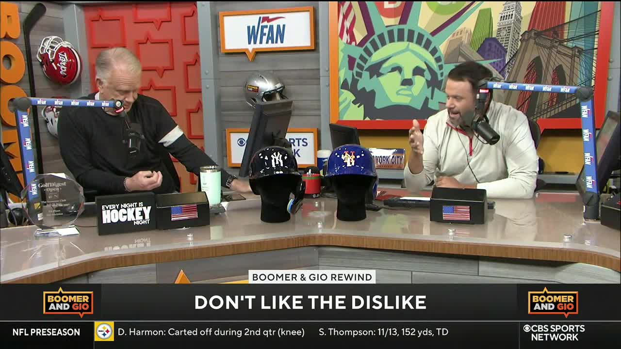 Two men sit behind a desk, discussing something on CBS Sports Network USA. Baseball helmets sit between them, and one man gestures with his hands as he speaks into a microphone.
