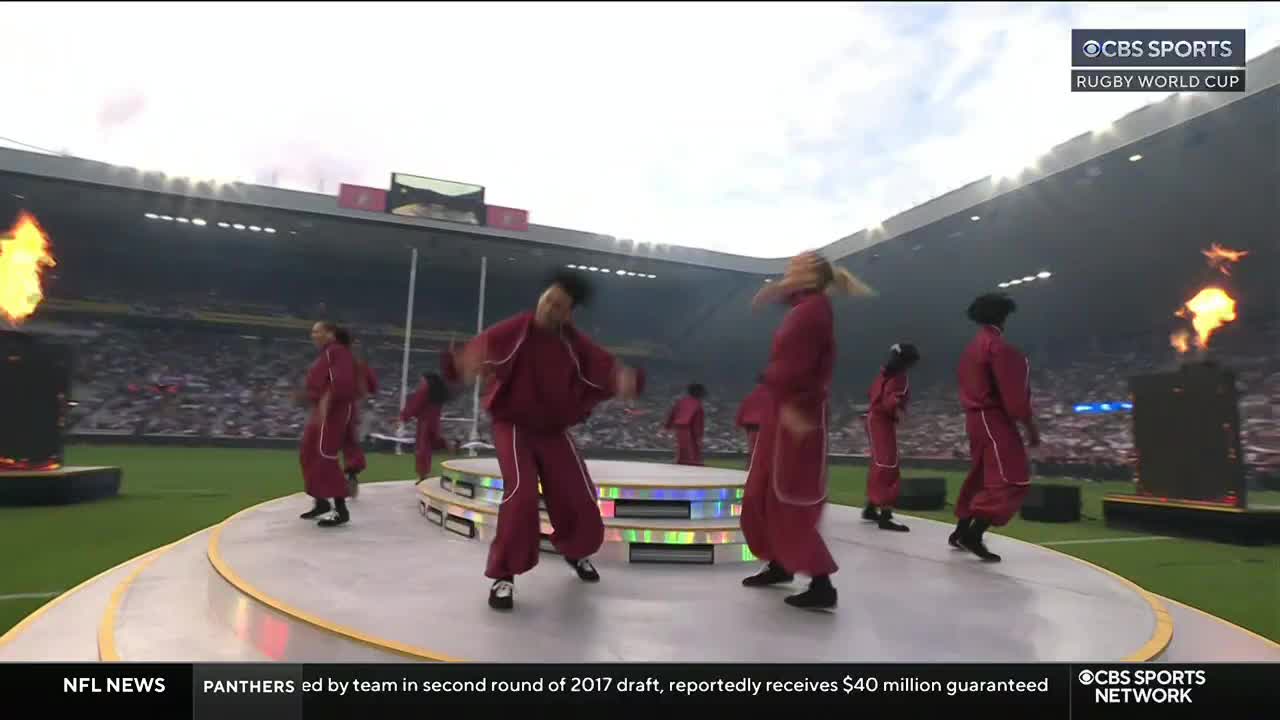 Dancers in maroon jumpsuits move on a raised platform, their arms and heads swaying.  A large flame burns to the left, and the CBS Sports Network logo is visible in the corner.

