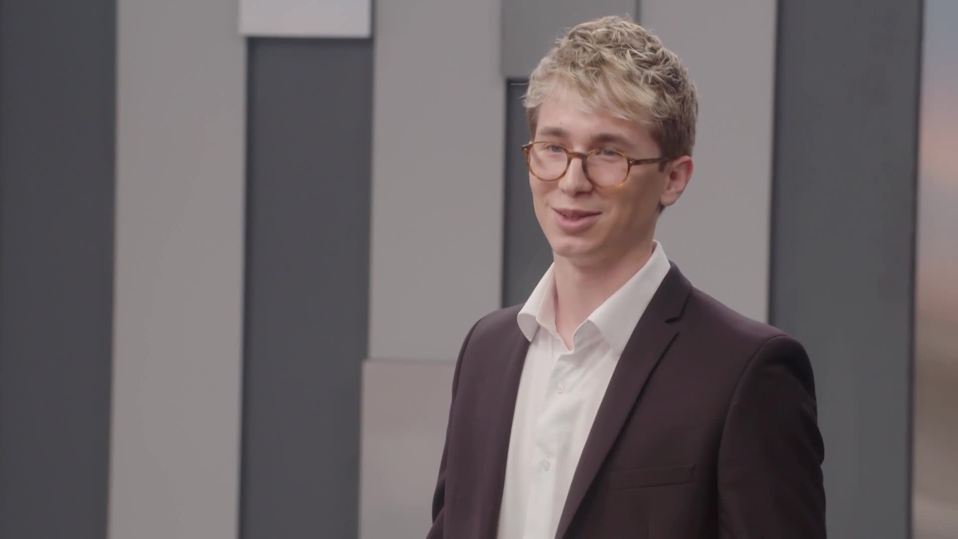 A young man in a suit and glasses stands in front of a gray and white striped backdrop. He's smiling, as if he's about to speak on Cheddar News.
