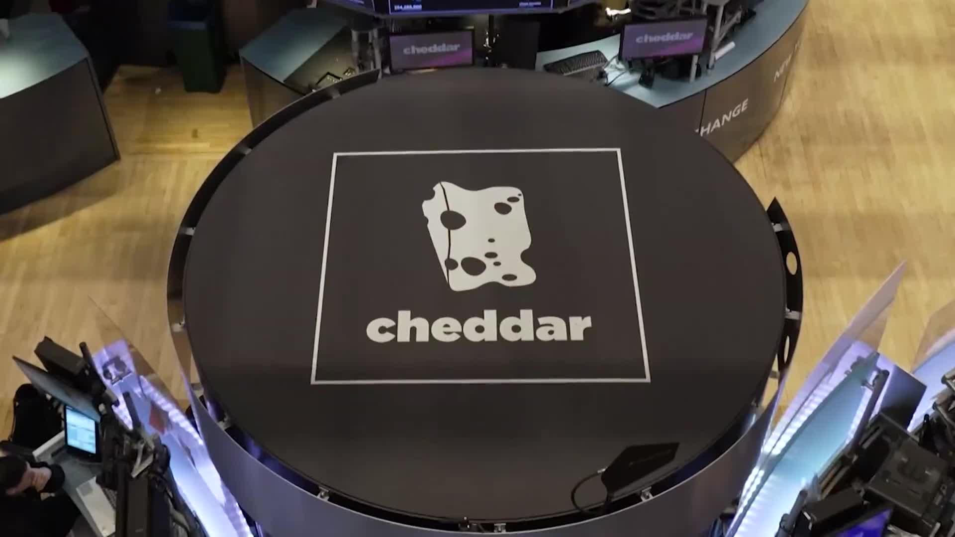 The Cheddar News logo, a square with a cheese illustration, is centered on a large black platform. Around it, people work at computer stations in a studio setting.
