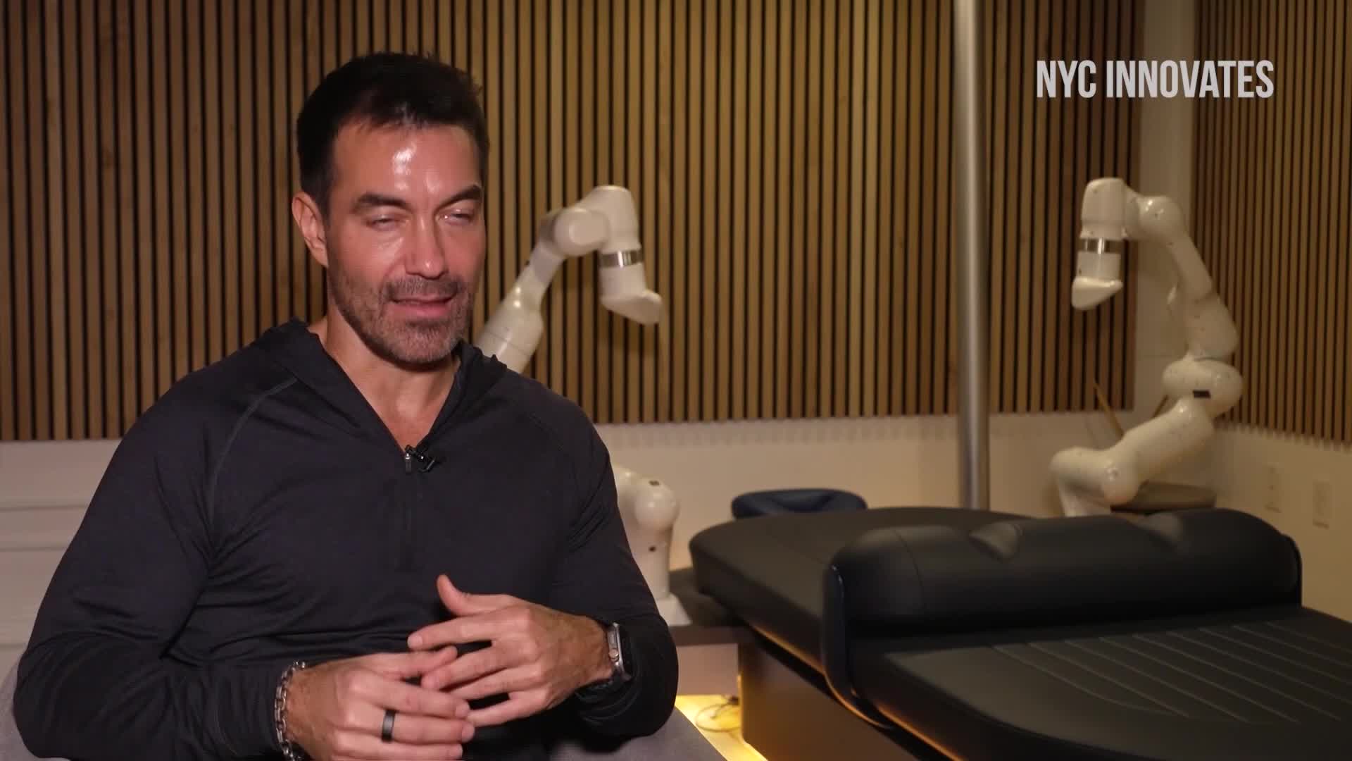 A man in a black shirt sits and speaks, while two white robotic arms hover near a massage table. The scene is likely part of a Cheddar News segment about innovation in the United States.

