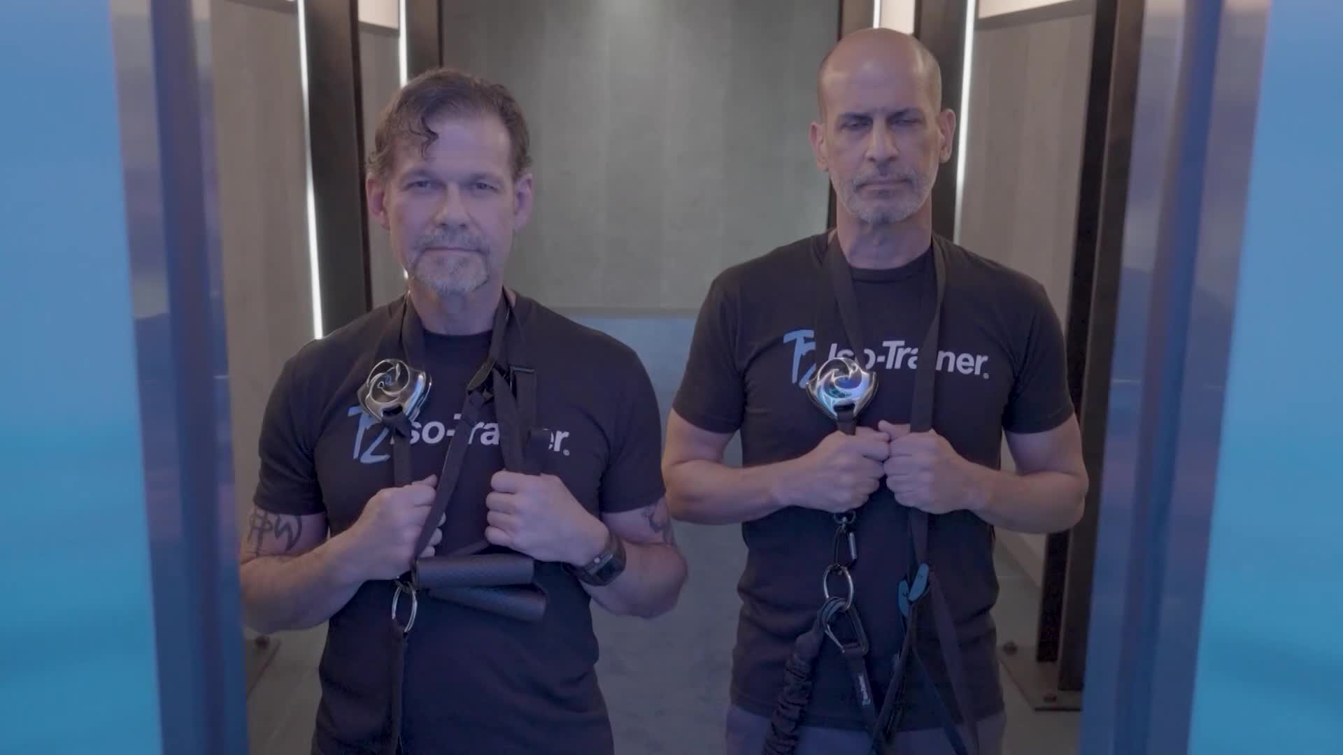 Two men, both wearing black t-shirts with "Iso-Trainer" printed on them, stand facing the camera. Each man holds the straps of a piece of exercise equipment, looking directly ahead.
