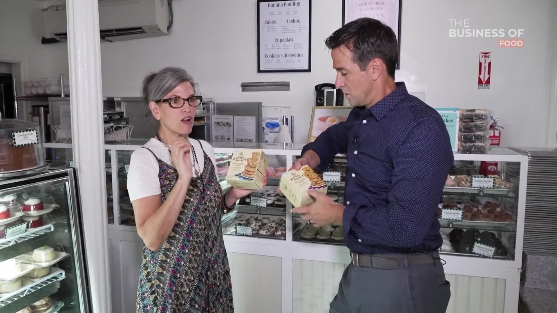 A woman in glasses gestures with one hand while holding a box of dessert. A man in a blue shirt holds another box of the same product, seemingly examining it.
