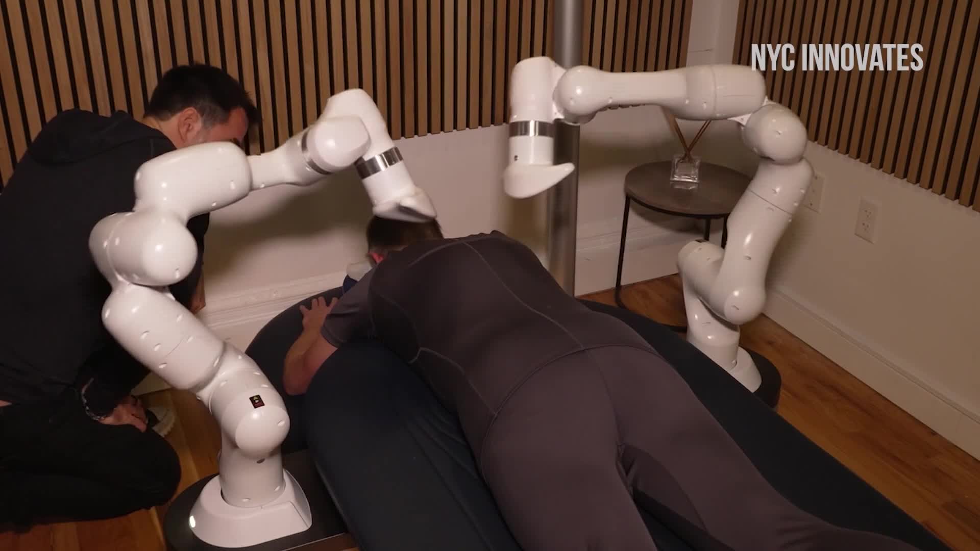 Two white robotic arms hover above a man lying face down on a massage table. A person stands beside the man, seemingly adjusting the robot's position.
