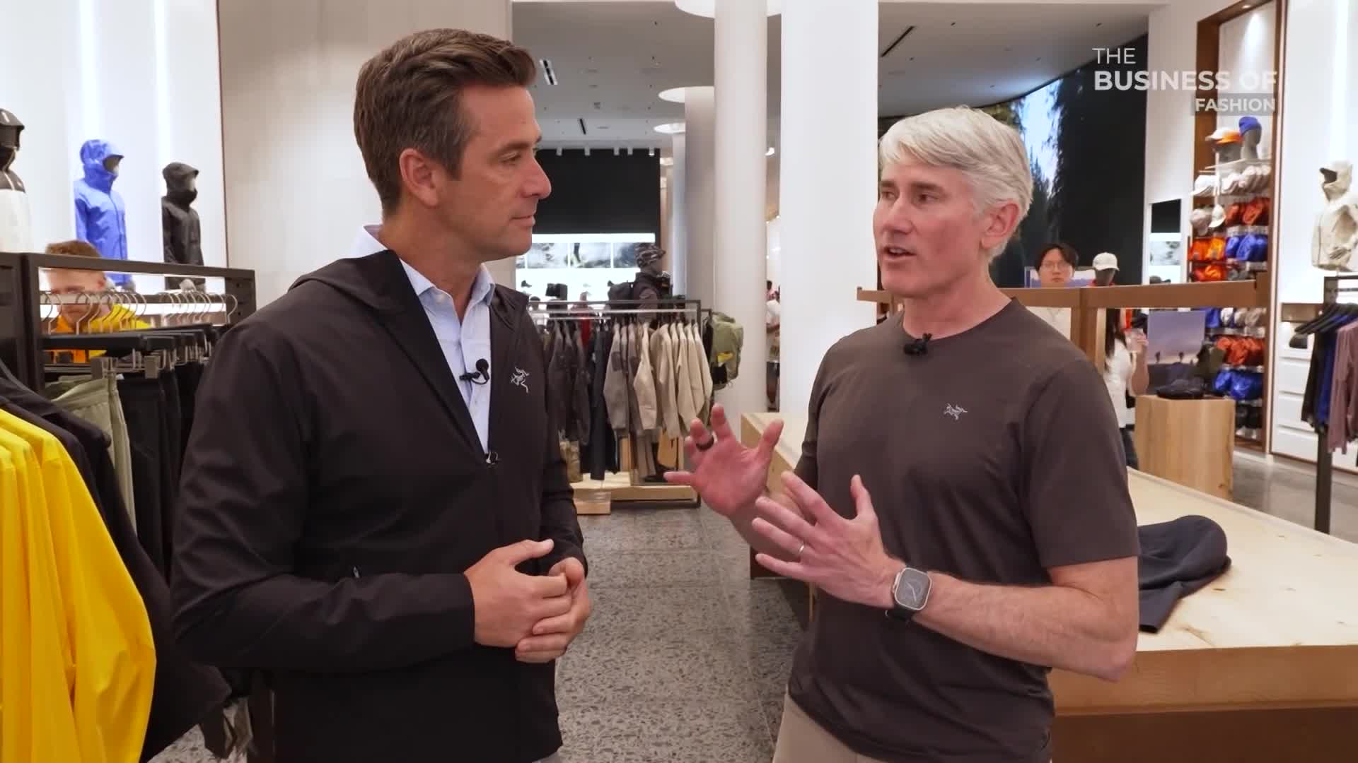 The man in the brown shirt gestures with his hands, speaking to the person in the black jacket. They're standing in a store, with racks of clothing visible in the background.
