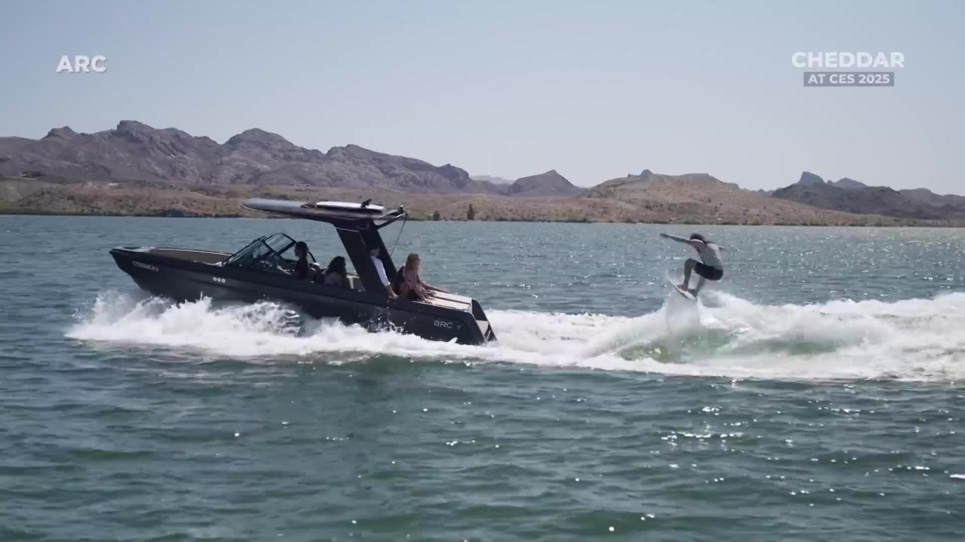 A sleek black boat cuts through the water, creating a wake for a surfer riding behind it. The person on the board balances with arms outstretched, enjoying the ride.
