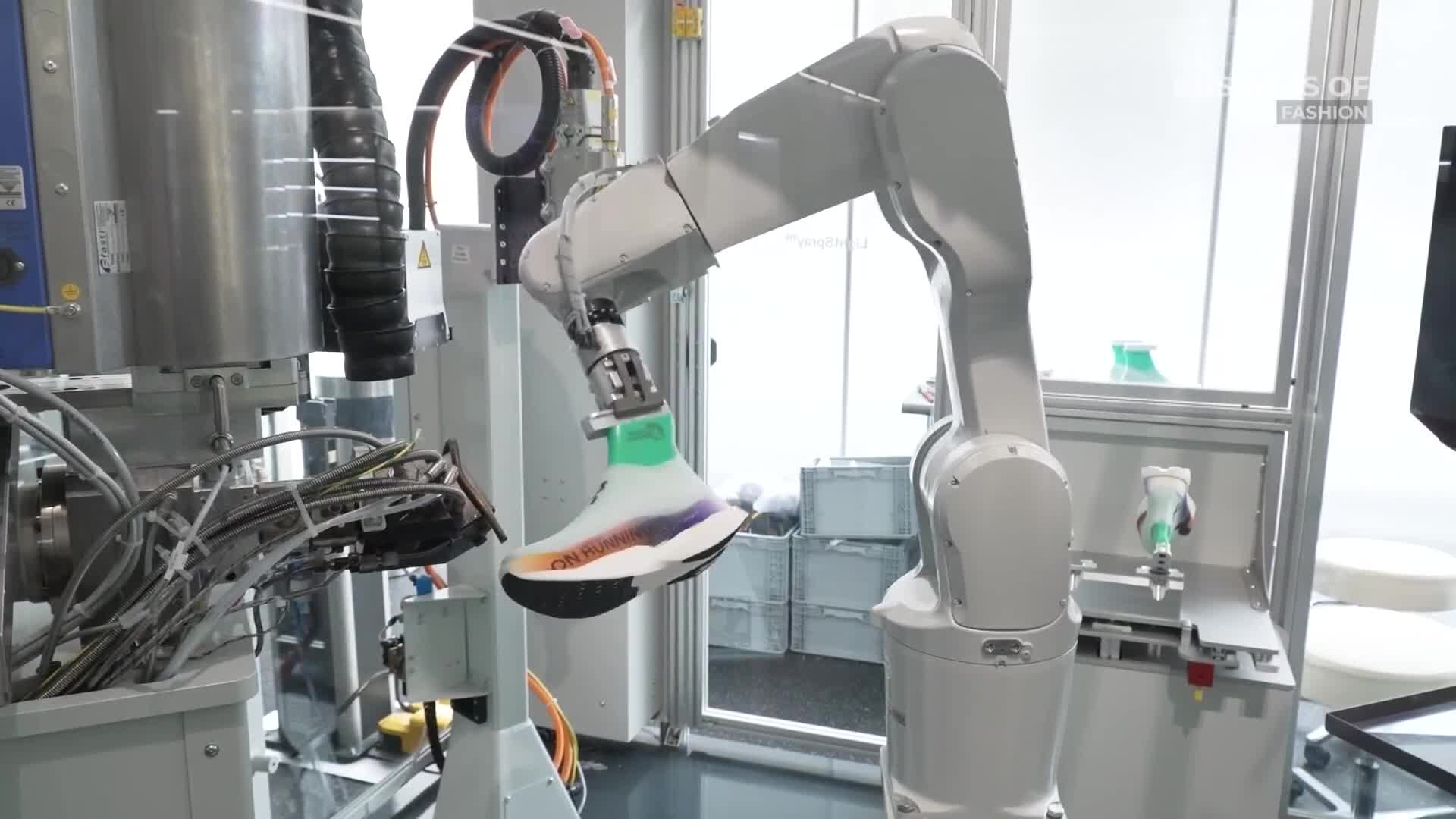 A robotic arm holds a sneaker, its grip secured around the shoe's upper. The arm maneuvers the shoe near a machine, likely for some stage of the manufacturing process.
