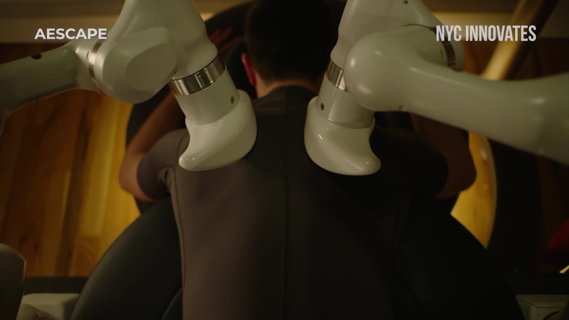 A person sits with their back to me, cradled by the curved, white arms of a robotic device. The scene is lit dimly, with the logos "AESCAPE" and "NYC INNOVATES" visible in the corners.
