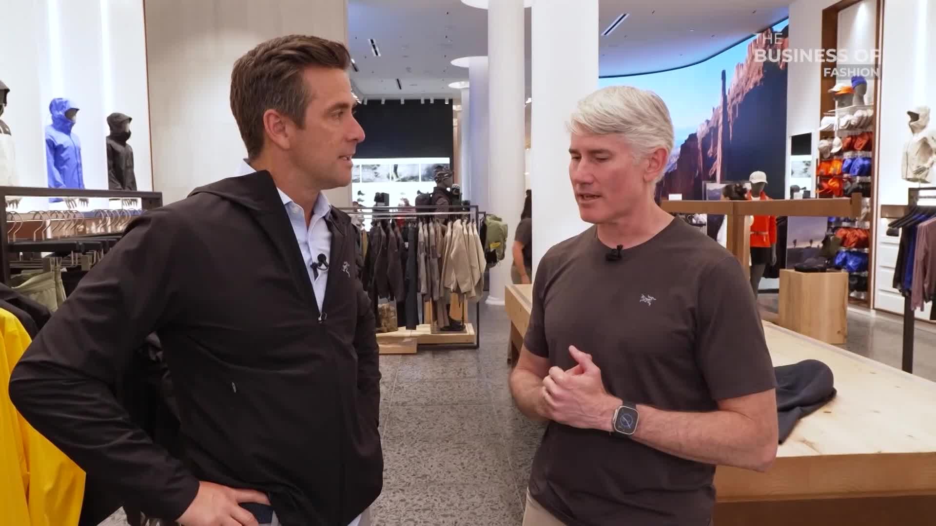 Two men are standing in a brightly lit retail space, seemingly engaged in a conversation. The man on the right gestures with his hands while the other listens, both wearing clothing branded with the same logo.
