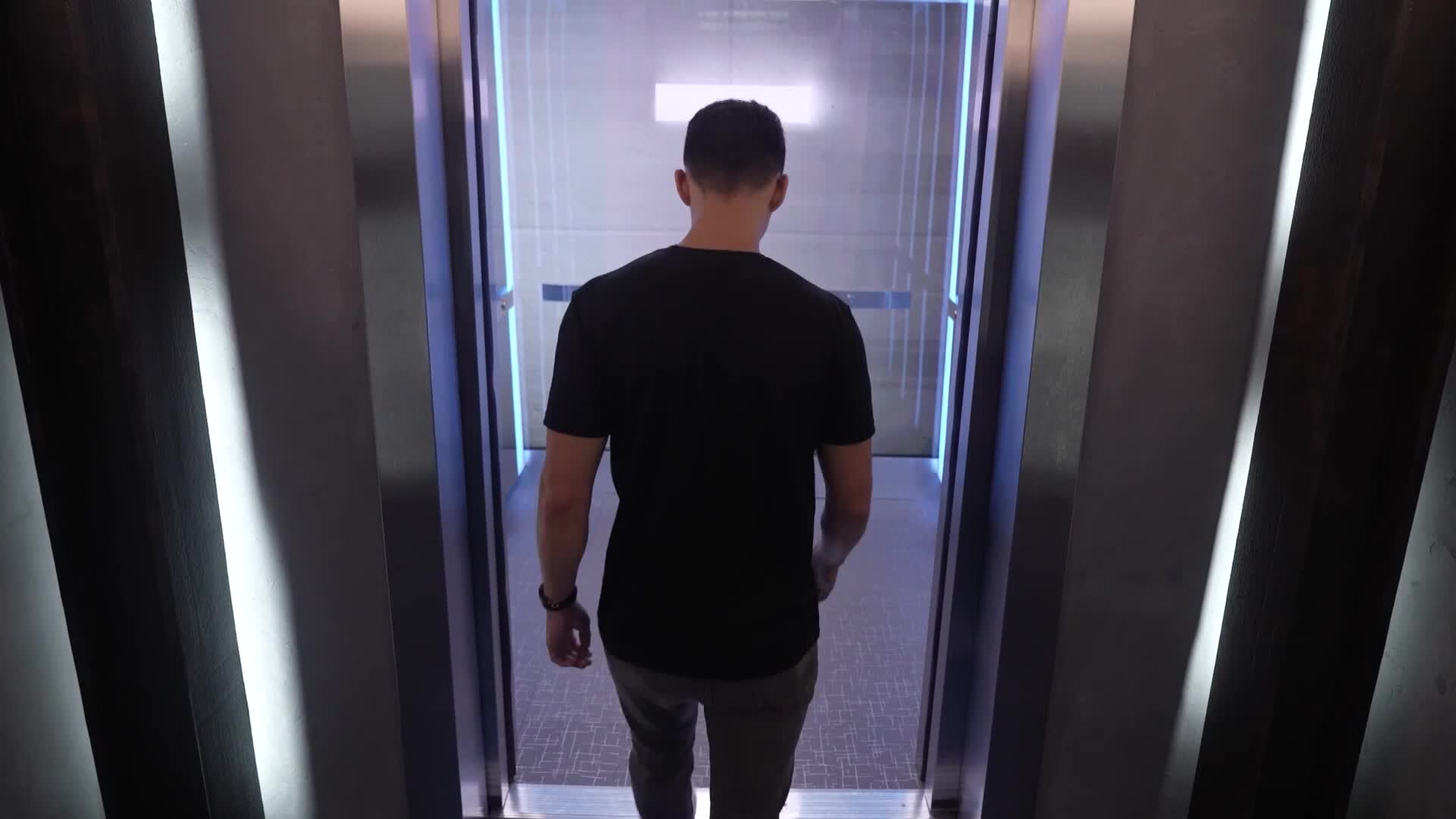 A man in a black t-shirt walks into an elevator, his back to the camera. The elevator doors are open, framed with blue neon lights.
