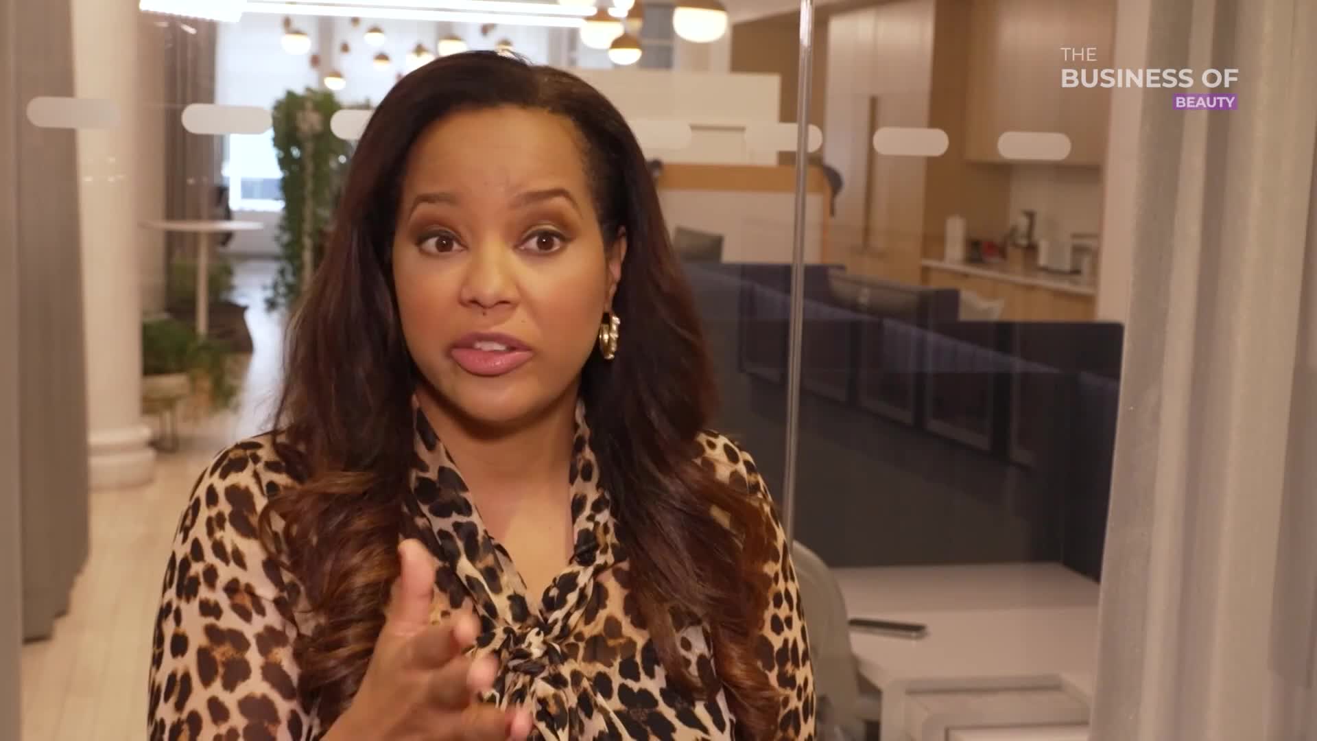 A woman in a leopard-print blouse gestures with her right hand as she speaks. The backdrop is a modern office space with glass walls and a "The Business of Beauty" logo.
