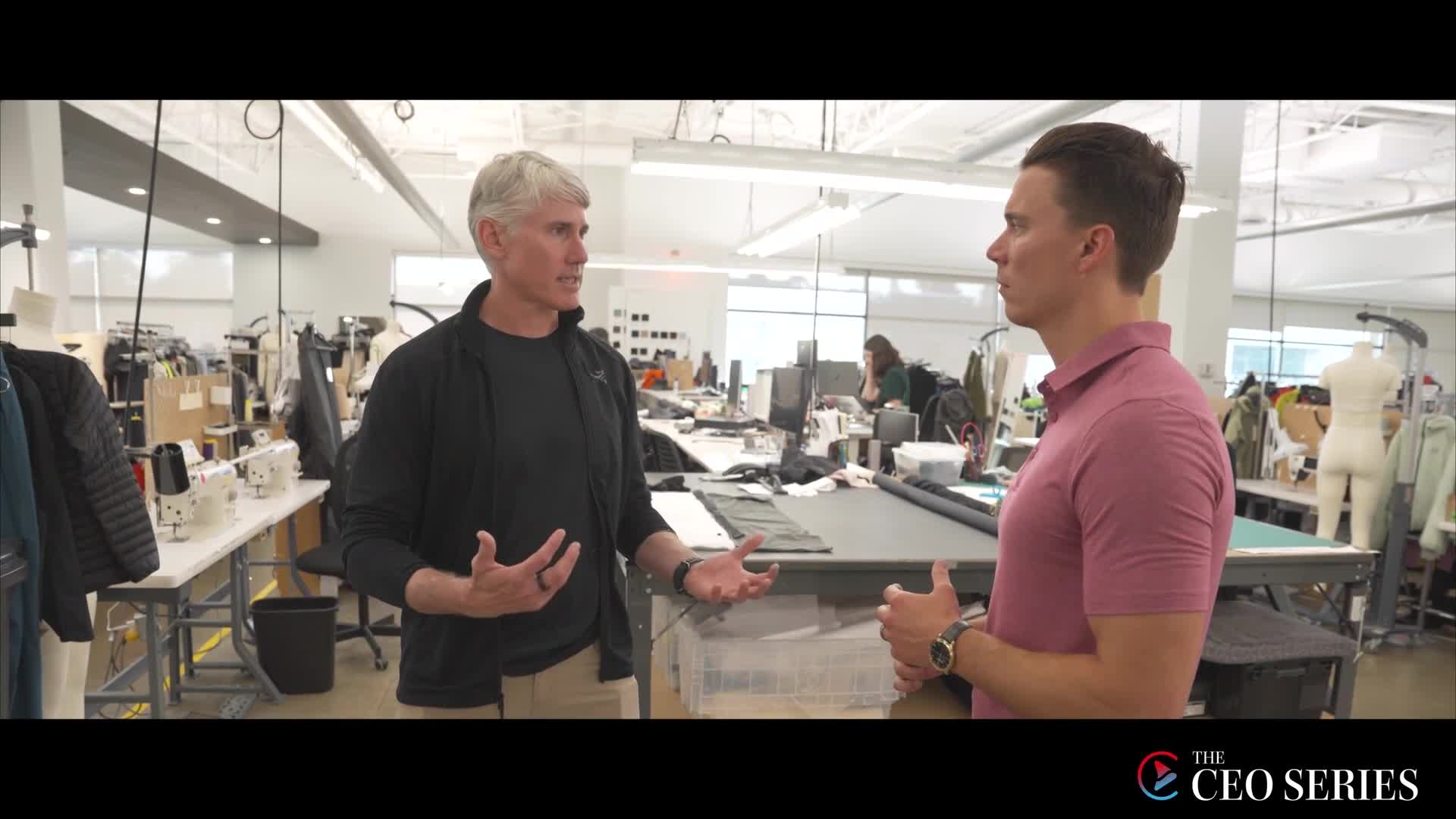 A man in a black jacket gestures with both hands, speaking to another man wearing a pink polo shirt. They're in a bright, open workshop filled with sewing machines and cutting tables.
