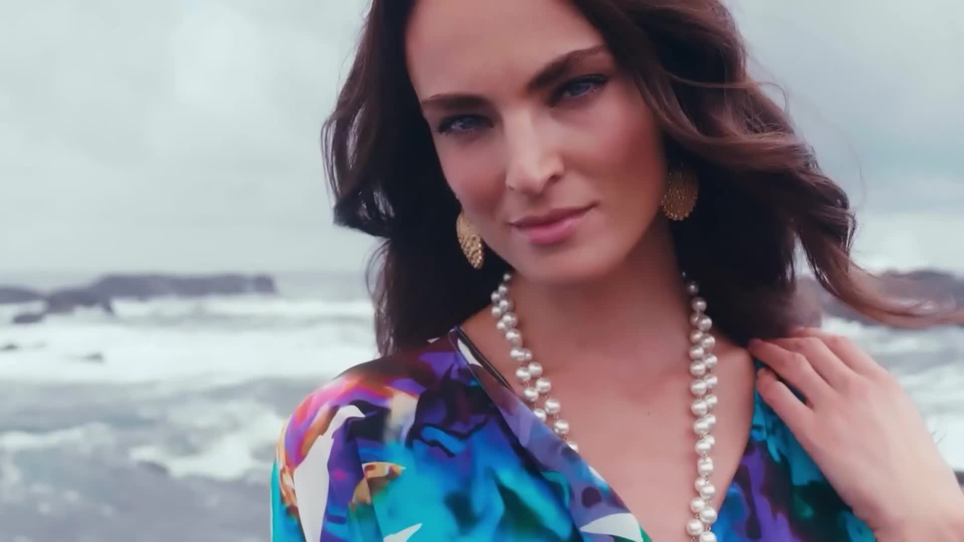 A woman with long, wavy hair and a pearl necklace looks directly at the camera, a colorful patterned top visible. The ocean is in the background, and the sky is overcast.
