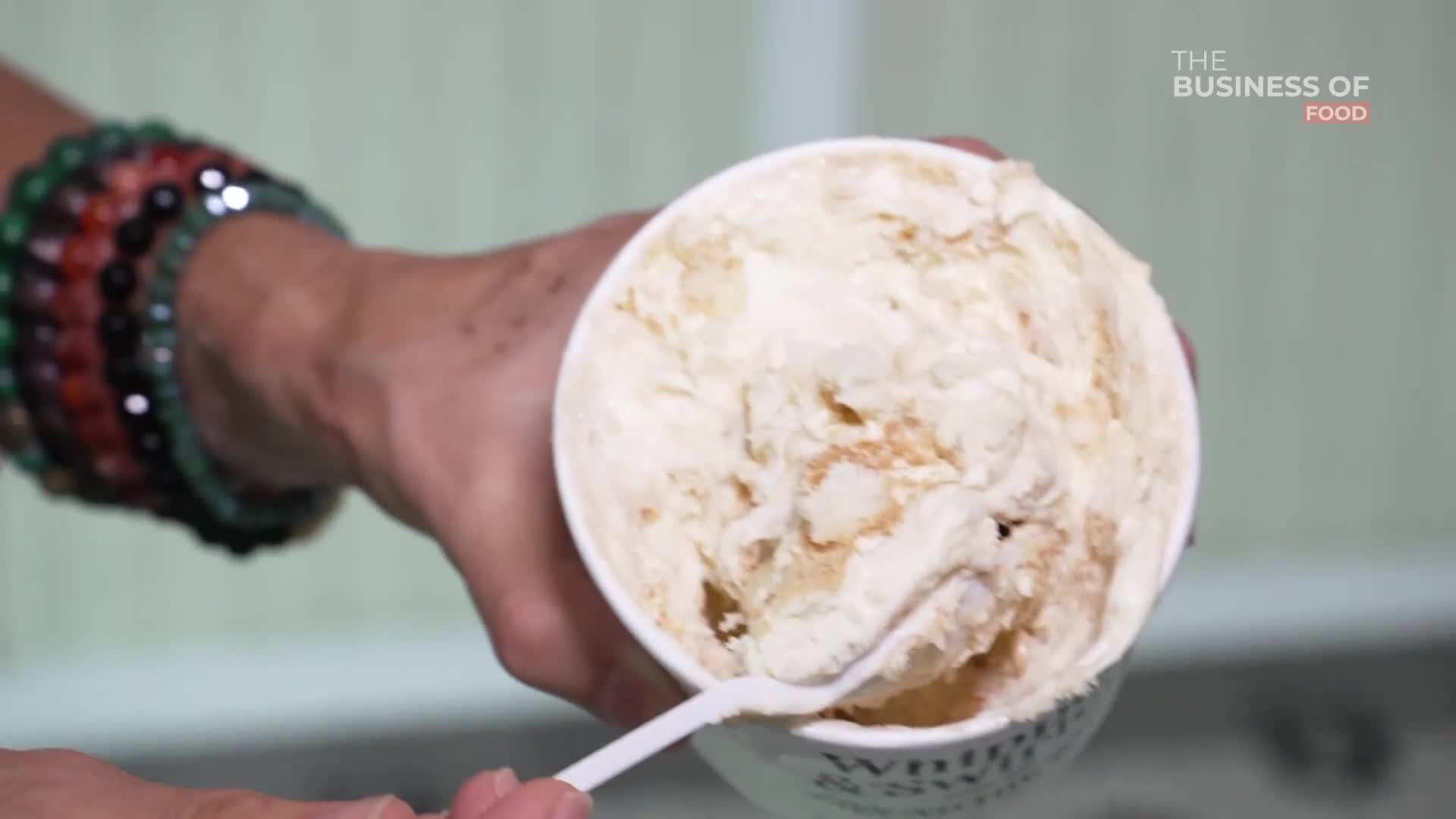 A hand, adorned with beaded bracelets, holds a container of ice cream. A white plastic spoon scoops up a portion of the creamy, light-brown dessert.
