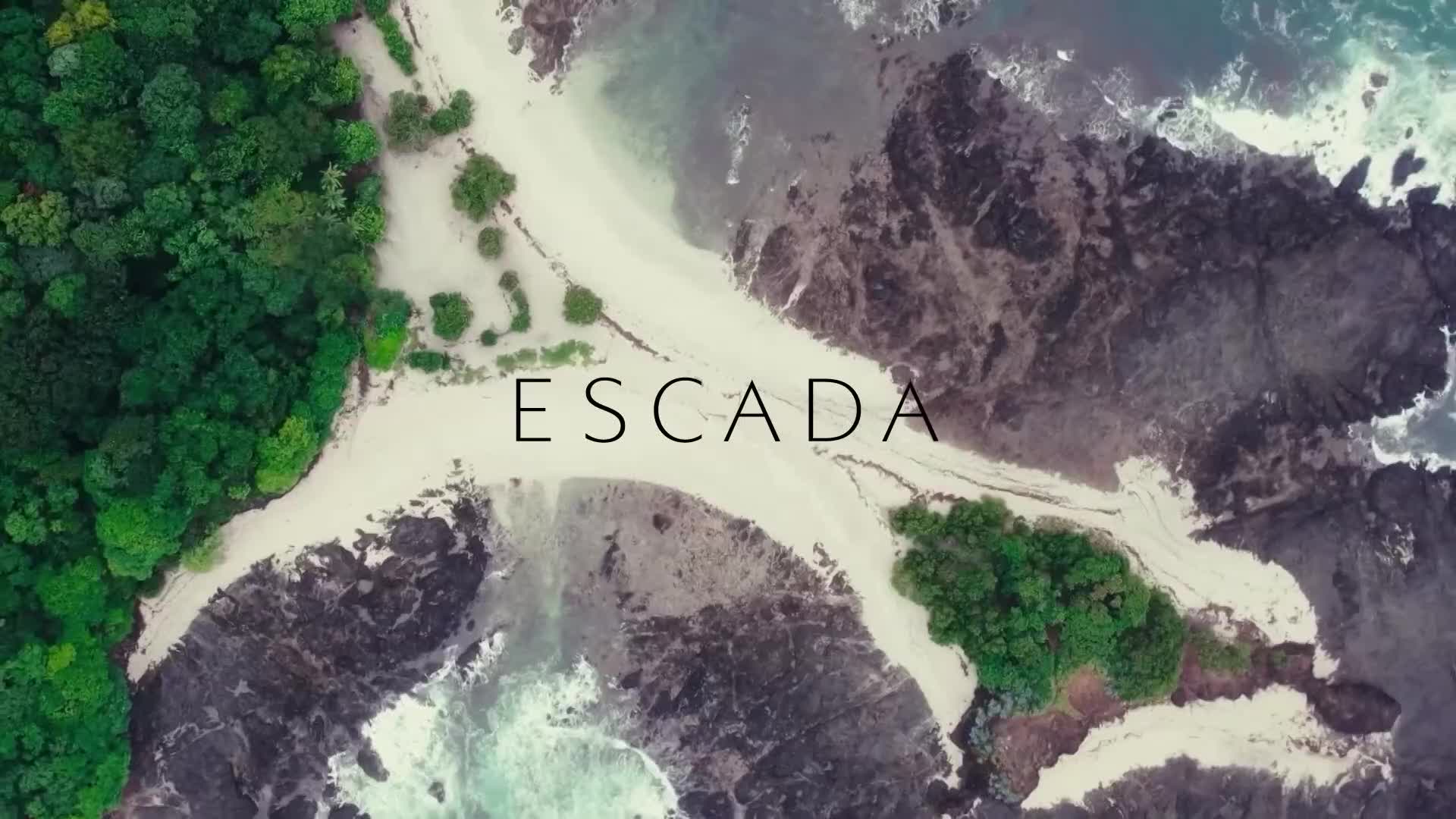 The word "ESCADA" floats above a sandy beach where the ocean meets the shore. Lush green trees border the white sand, and the dark rocks of the coastline jut into the water.
