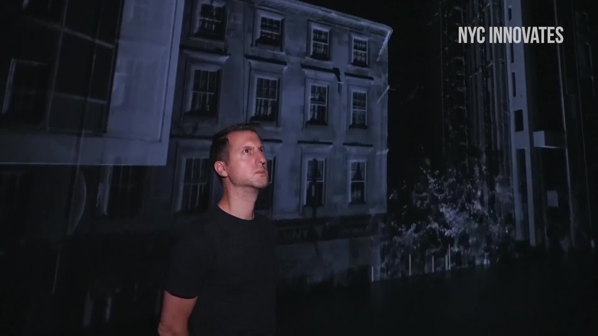 A man in a black t-shirt stands in front of a projected image of a building. The building appears to be part of a cityscape, possibly in the United States, and is illuminated in a dim light.
