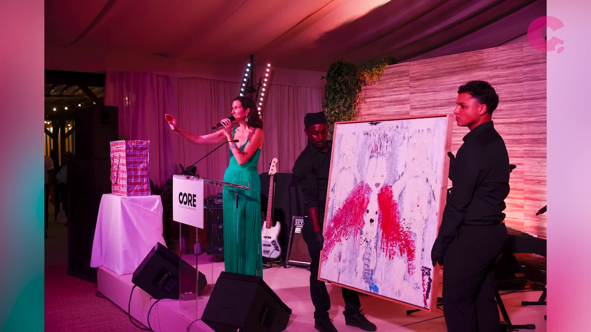 A woman in a green dress speaks into a microphone on a stage, gesturing with her right hand. Two men carry a large painting with red and white splatters across it.
