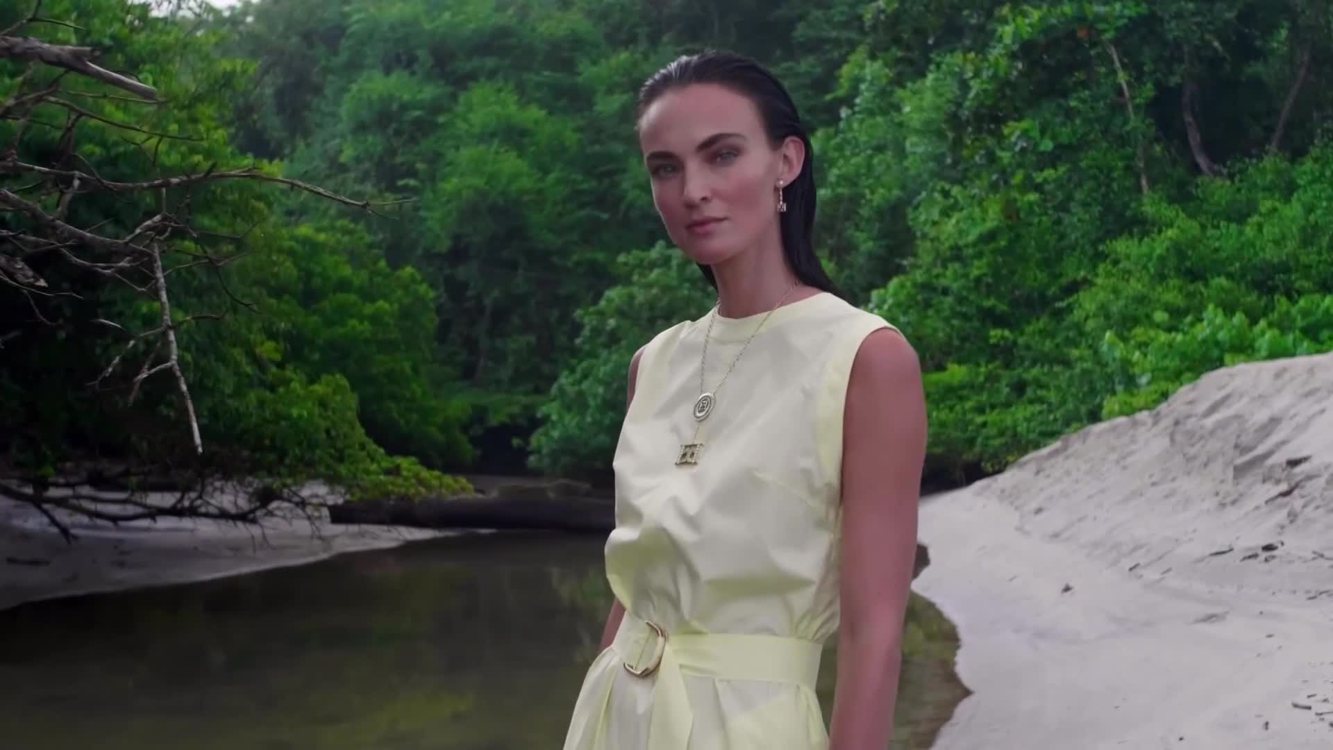 A woman in a pale yellow dress stands on a sandy beach, a small stream flowing behind her. Lush green foliage frames the scene, and the woman looks directly at the camera.
