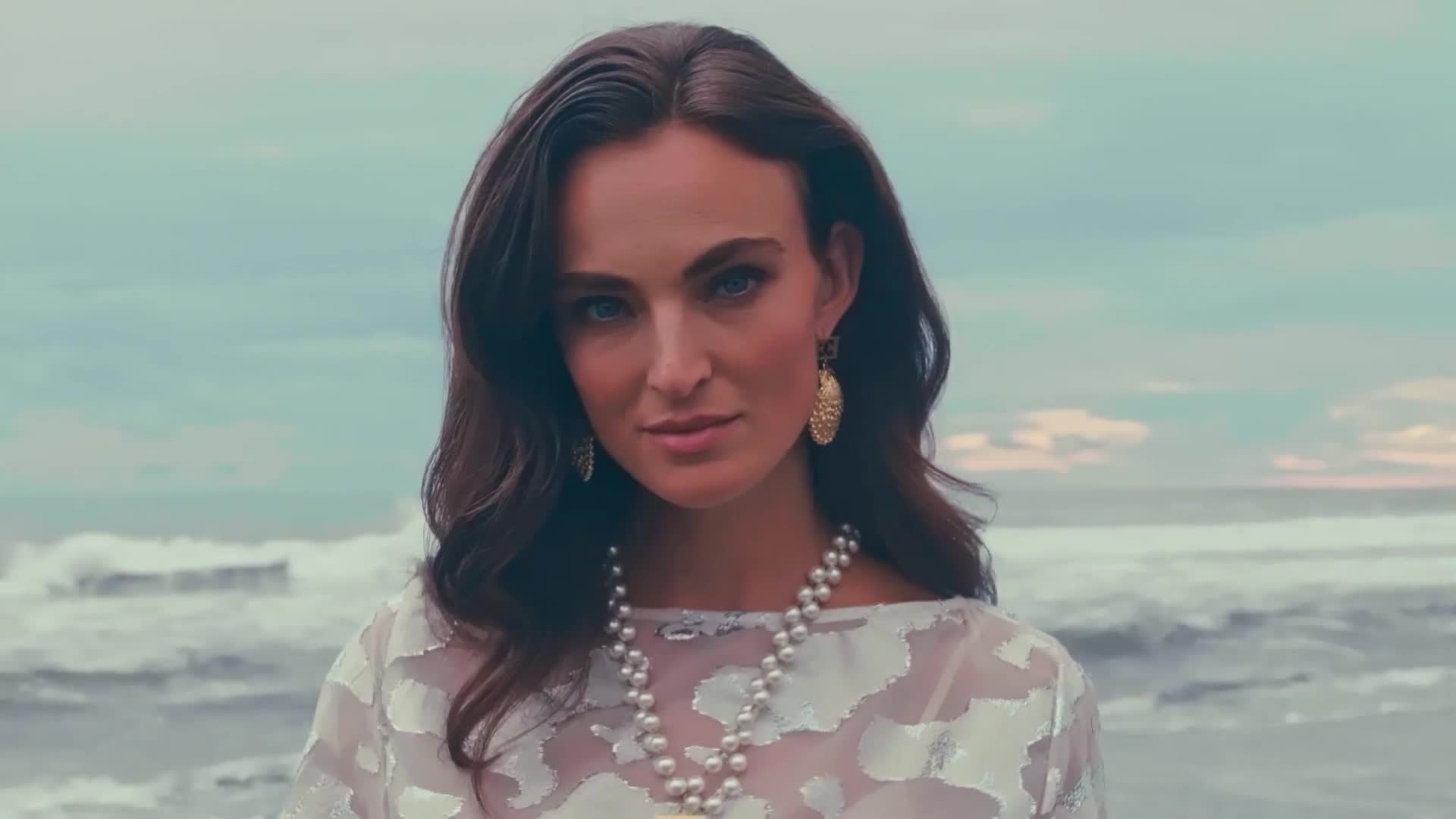A woman with dark hair and pearl necklaces gazes directly at the camera, the ocean visible behind her. The waves are crashing in the distance, and the sky is a muted blue.
