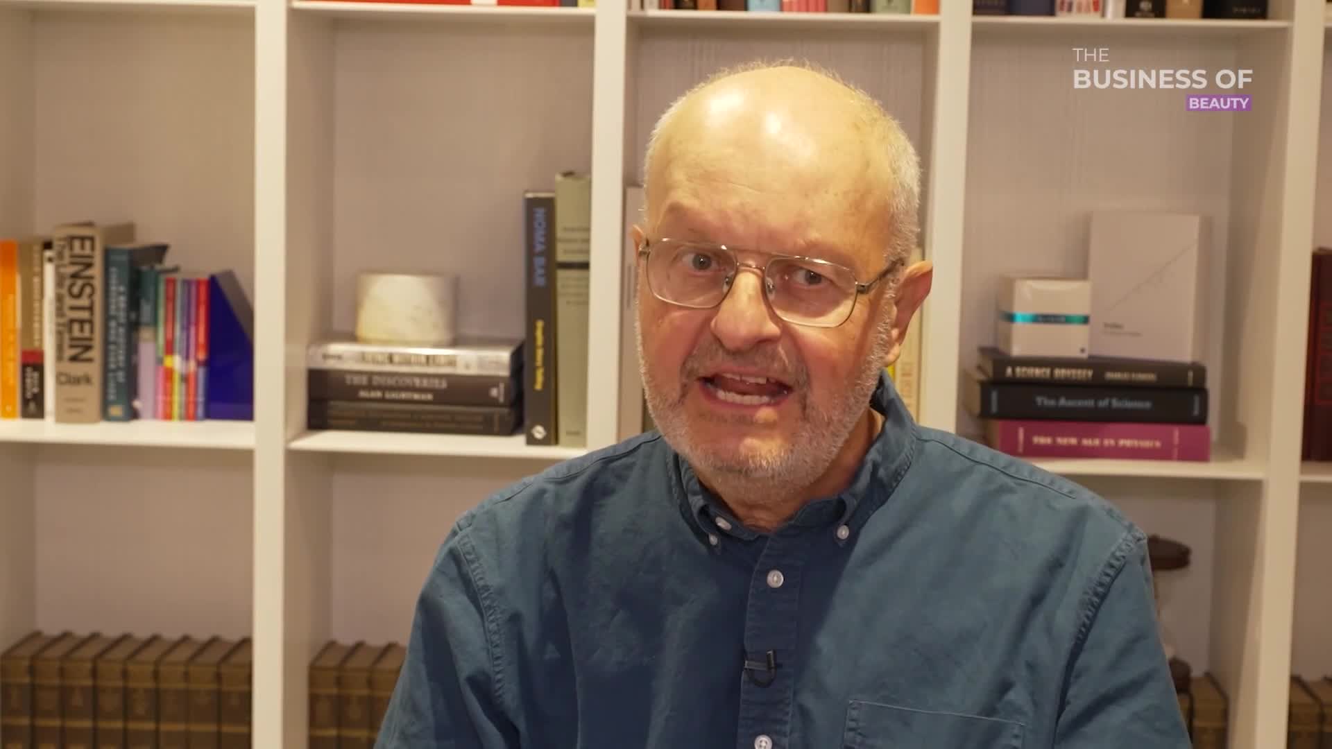 A man with glasses and a blue shirt is speaking, his mouth open in mid-sentence. Behind him, a white bookshelf is filled with books, and the "Business of Beauty" logo is visible in the corner.
