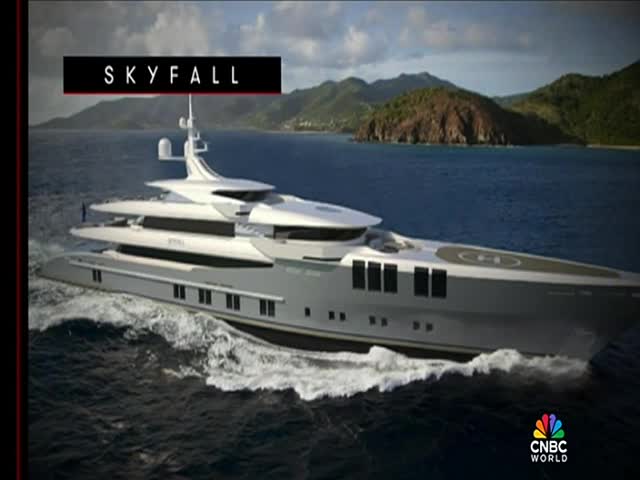 A sleek, white yacht cuts through the water, leaving a foamy wake. The vessel, labeled "SKYFALL" on the screen, appears on CNBC World, with a backdrop of green hills and blue sea.
