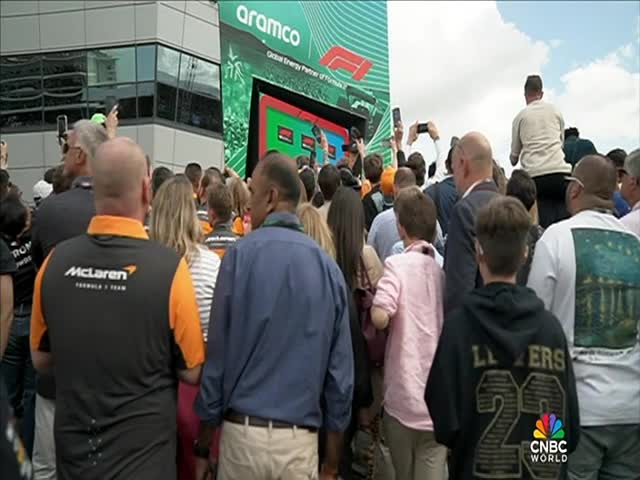 A crowd is gathered, many holding up phones, in front of a large screen with the Aramco logo. People in McLaren team shirts are interspersed throughout the throng.
