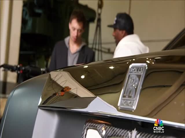 The front end of a Rolls-Royce gleams under the studio lights. Two men are chatting near the car, possibly discussing the CNBC World segment being filmed.
