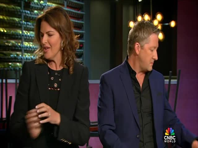The woman is gesturing with her hands, speaking with a slight smile. Beside her, the man in a blue jacket looks down, his right arm moving slightly.

