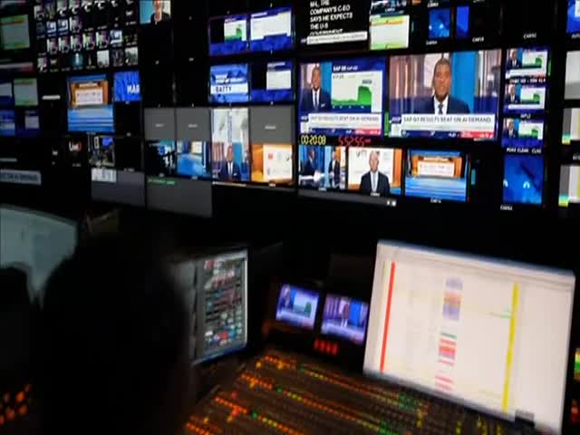 A wall of screens displays live news coverage, with CNBC World anchors reporting on the day's financial news. A technician's console is lit up, with a computer screen showing a detailed program schedule.
