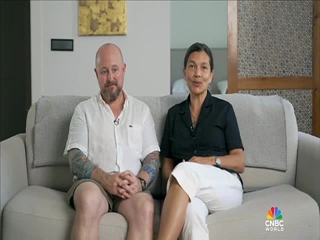 A man and a woman sit side-by-side on a gray couch, facing the camera. Both are wearing microphones, and the CNBC World logo is visible in the corner.
