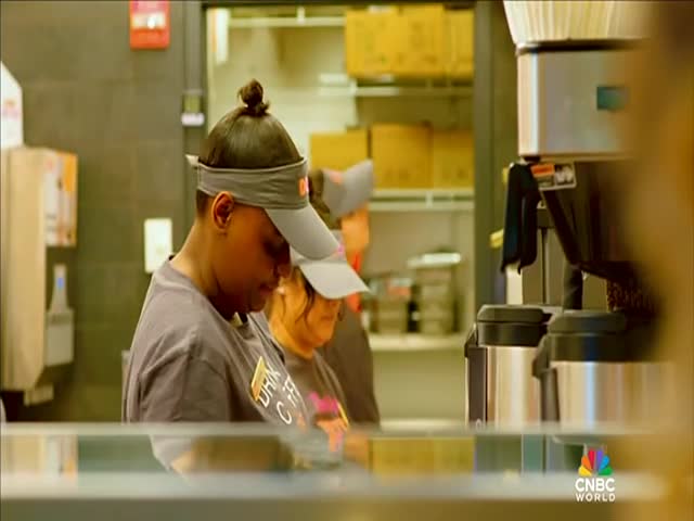 Inside, two employees in gray shirts and visors work behind a counter. One is focused on something out of frame, while the other is partially obscured.
